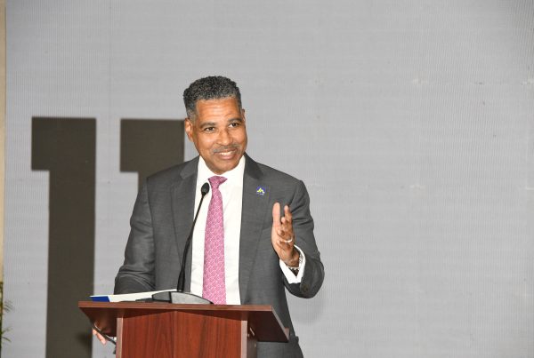 Man in a gray suit and pink tie speaks at a wooden podium with a microphone, gesturing with his right hand at a formal event.