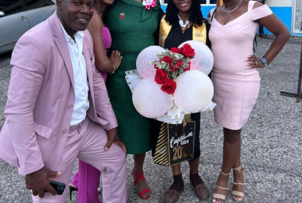 Five people pose together outside a building; a graduate in a cap and gown holds balloons and roses while others smile beside her.