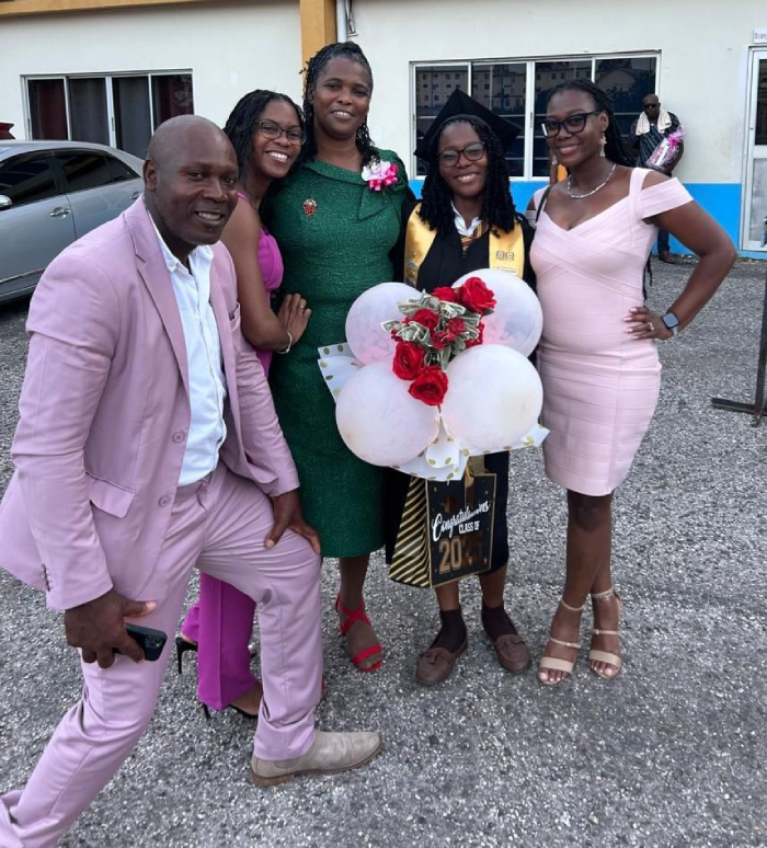 Five people pose together outside a building; a graduate in a cap and gown holds balloons and roses while others smile beside her.
