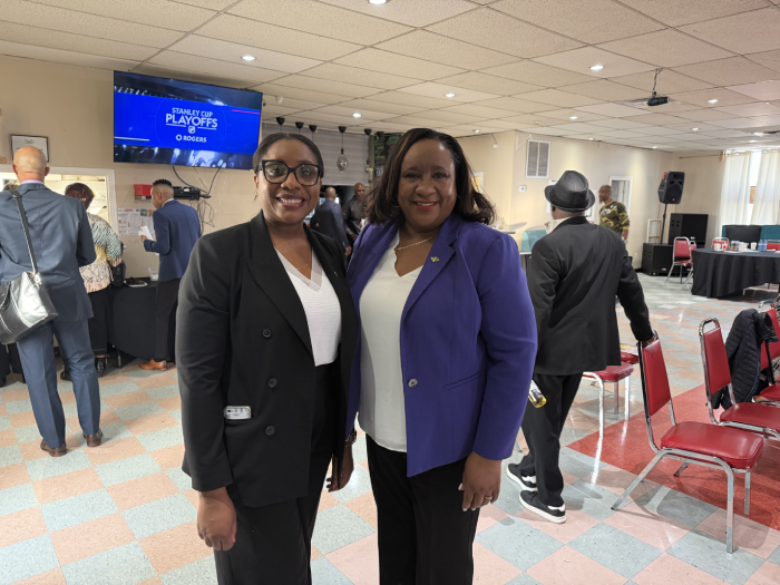 Two smiling women in black and purple blazers pose for a photo at an indoor event, with others in the background and a screen showing 'Stanley Cup Playoffs'.