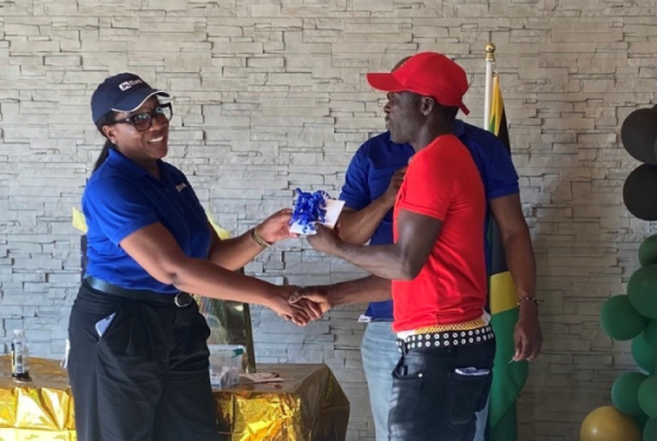 Two people shake hands during a ceremony: a woman in a blue polo hands a wrapped gift to a man in a red shirt, with gold table decorations and a flag in the background.