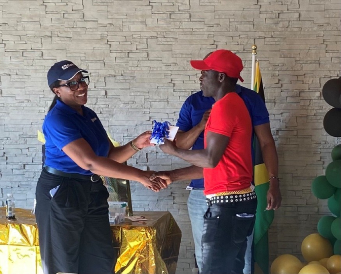 Two people shake hands during a ceremony: a woman in a blue polo hands a wrapped gift to a man in a red shirt, with gold table decorations and a flag in the background.