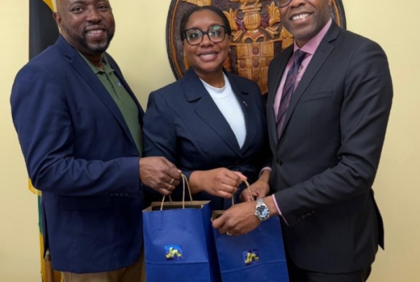 Three professionals in suits smile while holding blue gift bags, standing in front of a crest and flag.