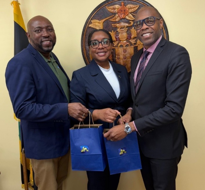 Three professionals in suits smile while holding blue gift bags, standing in front of a crest and flag.