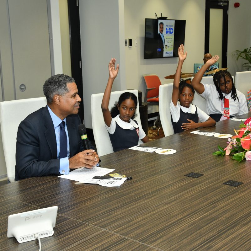 Man in a suit speaks into a microphone at a conference table as four young girls raise their hands in a classroom-style panel, with a flower arrangement in the foreground and banners in the background.