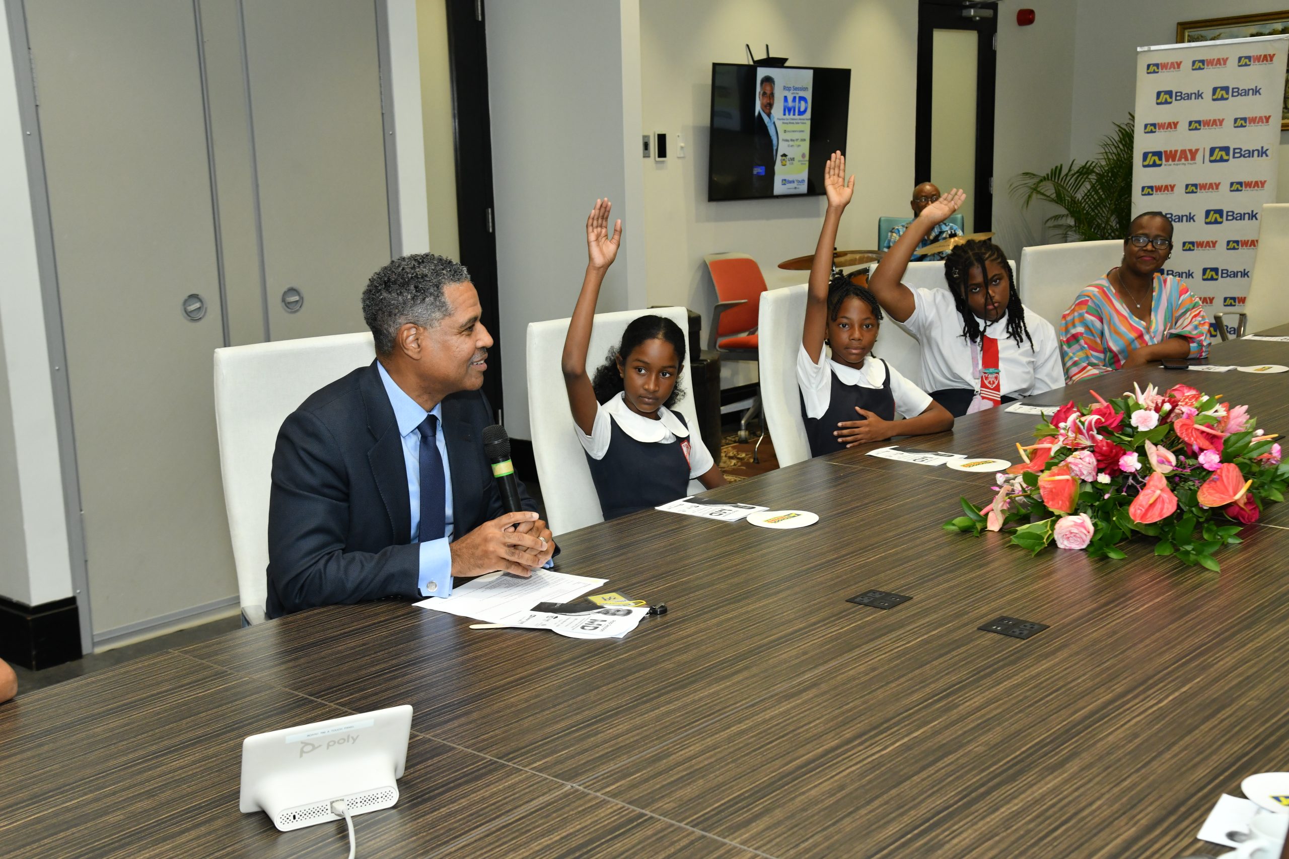 Man in a suit speaks into a microphone at a conference table as four young girls raise their hands in a classroom-style panel, with a flower arrangement in the foreground and banners in the background.