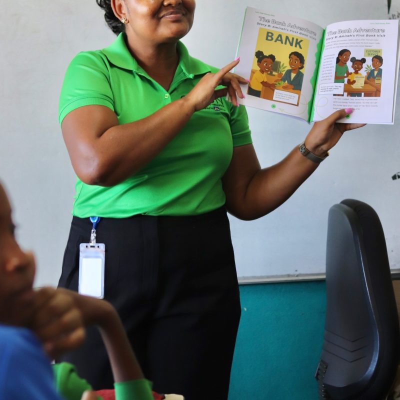 Woman in a bright green polo holds up an illustrated children's book about a bank, presenting to students in a classroom.
