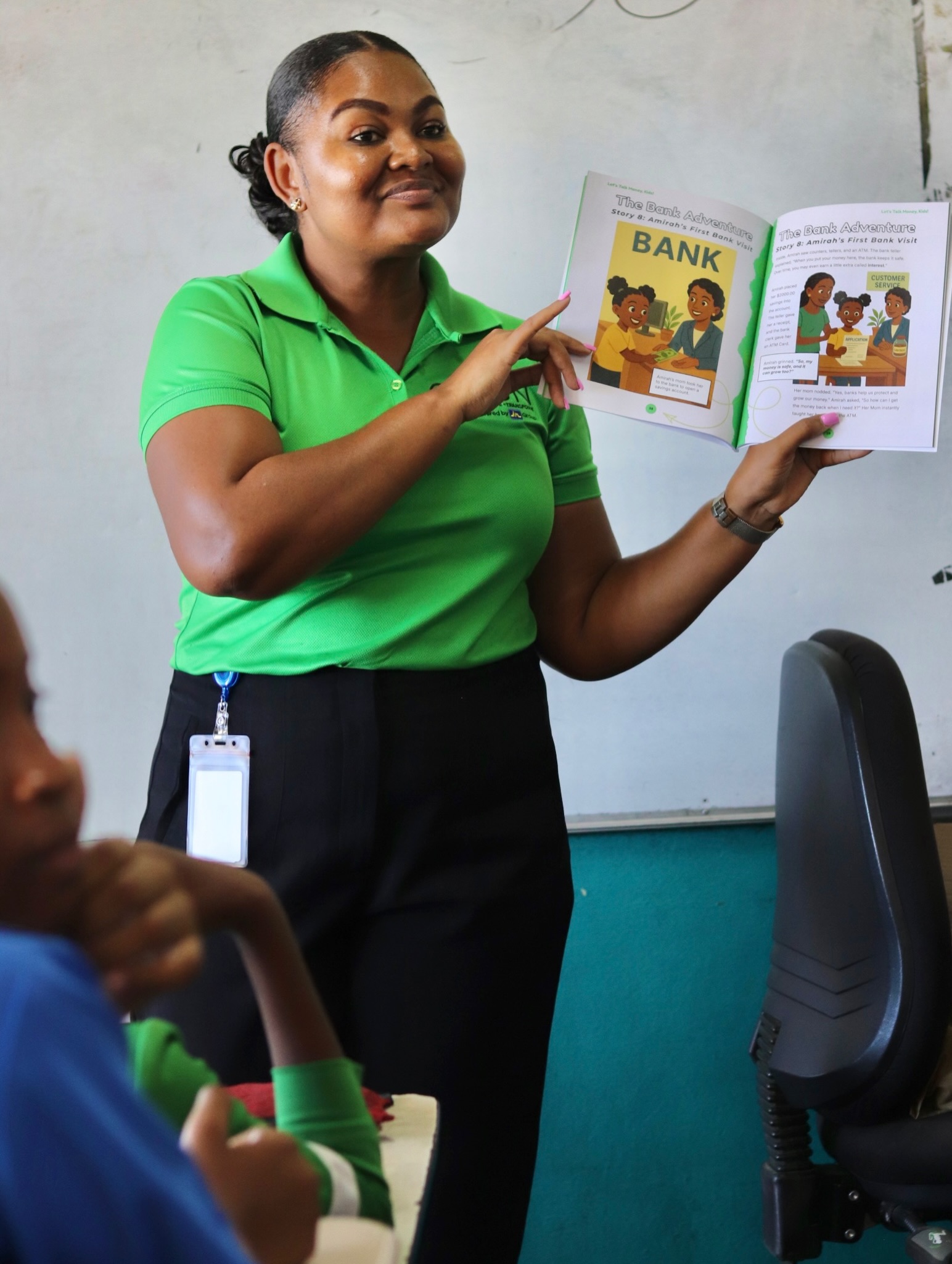 Woman in a bright green polo holds up an illustrated children's book about a bank, presenting to students in a classroom.