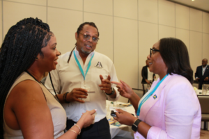 Three professionals converse at a conference, with two women and a man wearing name badges and teal lanyards, standing by a table with materials.