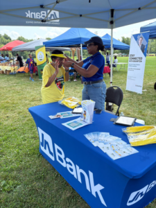 Two women at a Brampton outreach booth under blue canopies at an outdoor community fair; a volunteer in a blue shirt helps a woman in a yellow shirt with a headband, standing beside a table covered with a blue cloth and brochures.