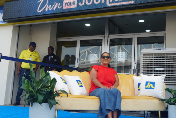 Woman in red blouse and sunglasses sits on a yellow sofa during a ribbon-cutting outside a store with an 'Own Your Journey' sign above the entrance.