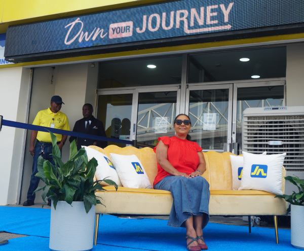 Woman in red blouse and sunglasses sits on a yellow sofa during a ribbon-cutting outside a store with an 'Own Your Journey' sign above the entrance.