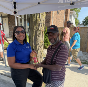 Woman in a blue polo hands a bottled drink to a man in a striped polo under a white canopy at a community event; both smiling, with other volunteers walking in the background.