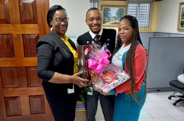 Three coworkers in an office pose while presenting a gift basket wrapped in pink ribbon; a man in a suit stands center flanked by two women.